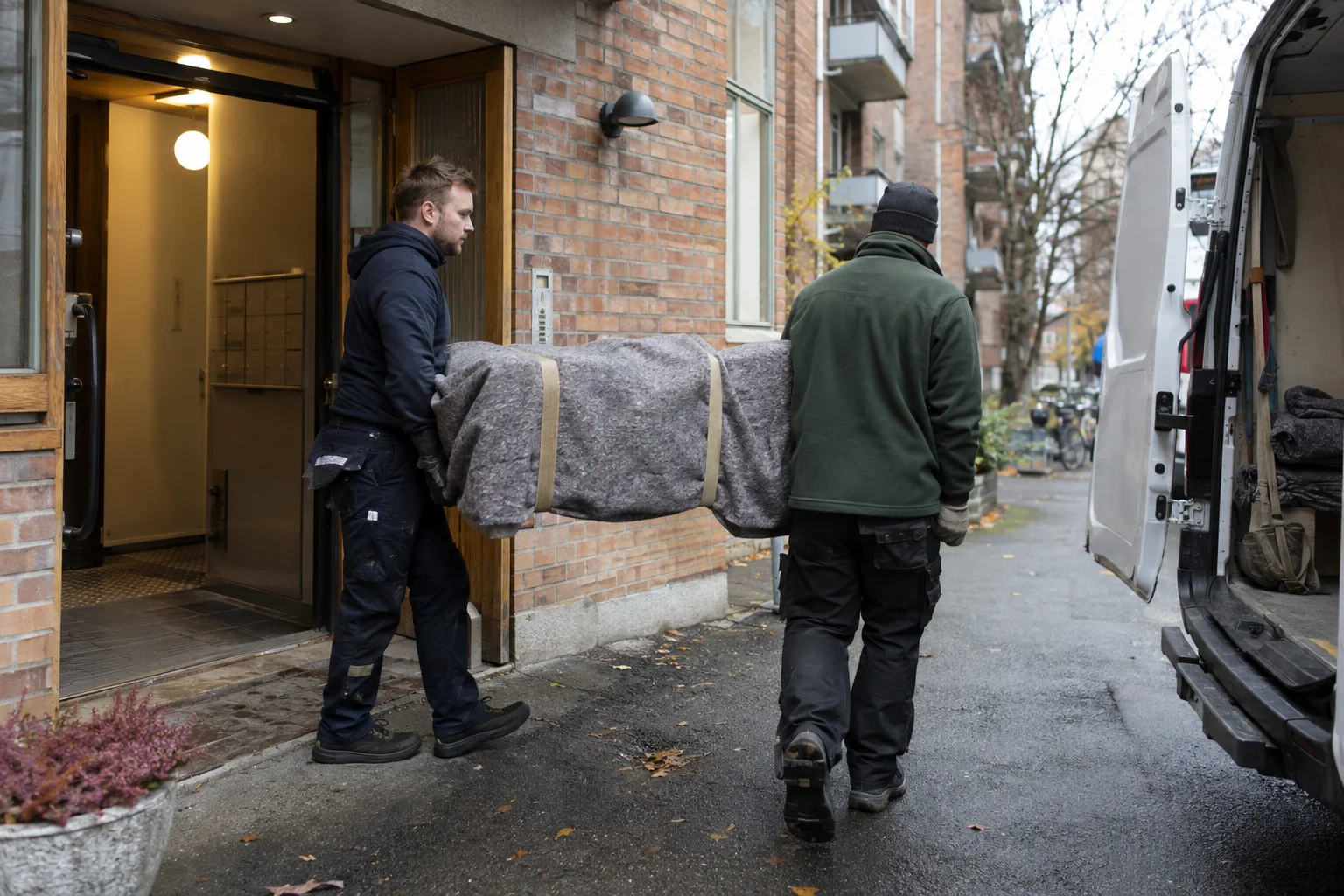 To flyttemedarbeidere bærer et beskyttet møbel fra en bygård mot en varebil